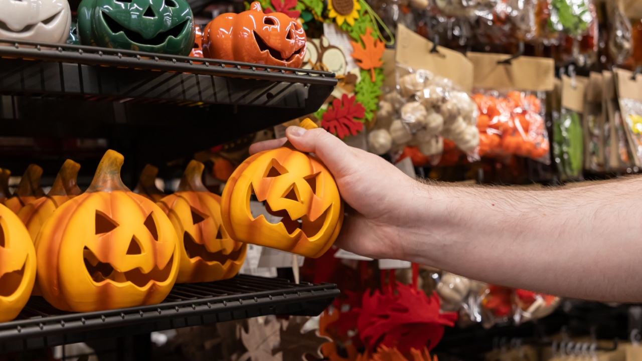 A detailed close-up view of a person's hand grasping an orange ceramic jack-o'-lantern, surrounded by seasonal Halloween merchandise on a bustling retail store shelf