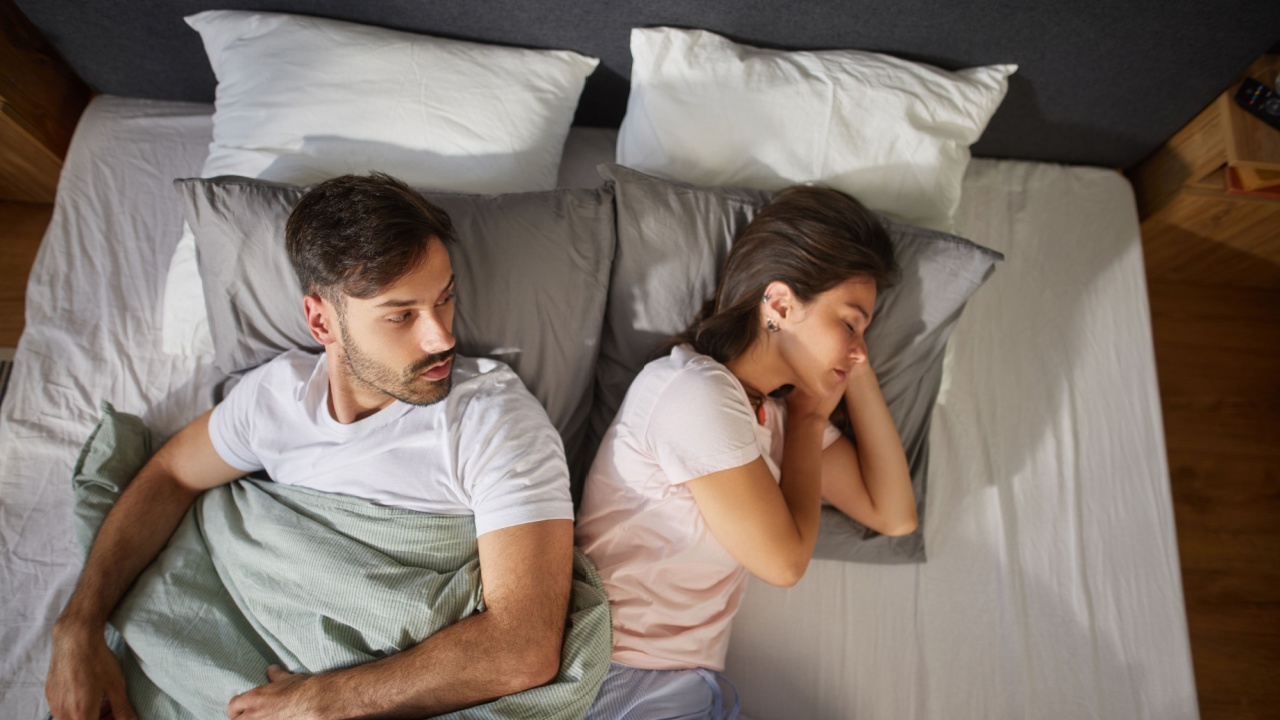 A couple lies in bed with their backs turned to one another, indicating emotional distance and unresolved relationship issues. Their expressions reveal discomfort and disconnect.