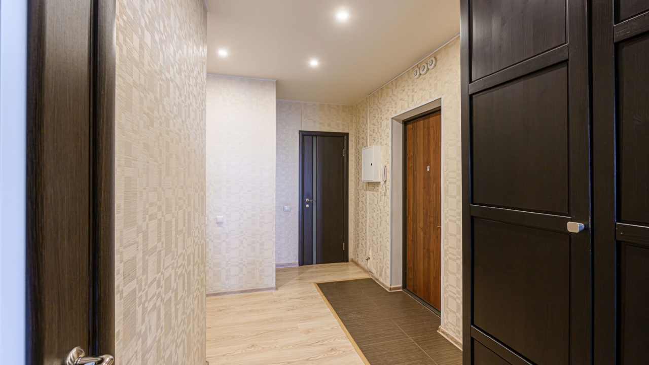 Hallway with patterned wallpaper, wood-look floor, and several doors. Dark wood wardrobe on the right. Recessed lighting