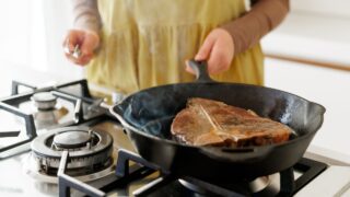Woman searing a T-bone steak in a cast iron skillet on a gas stove, with steam rising, perfect for cooking, grilling, and gourmet food themes.