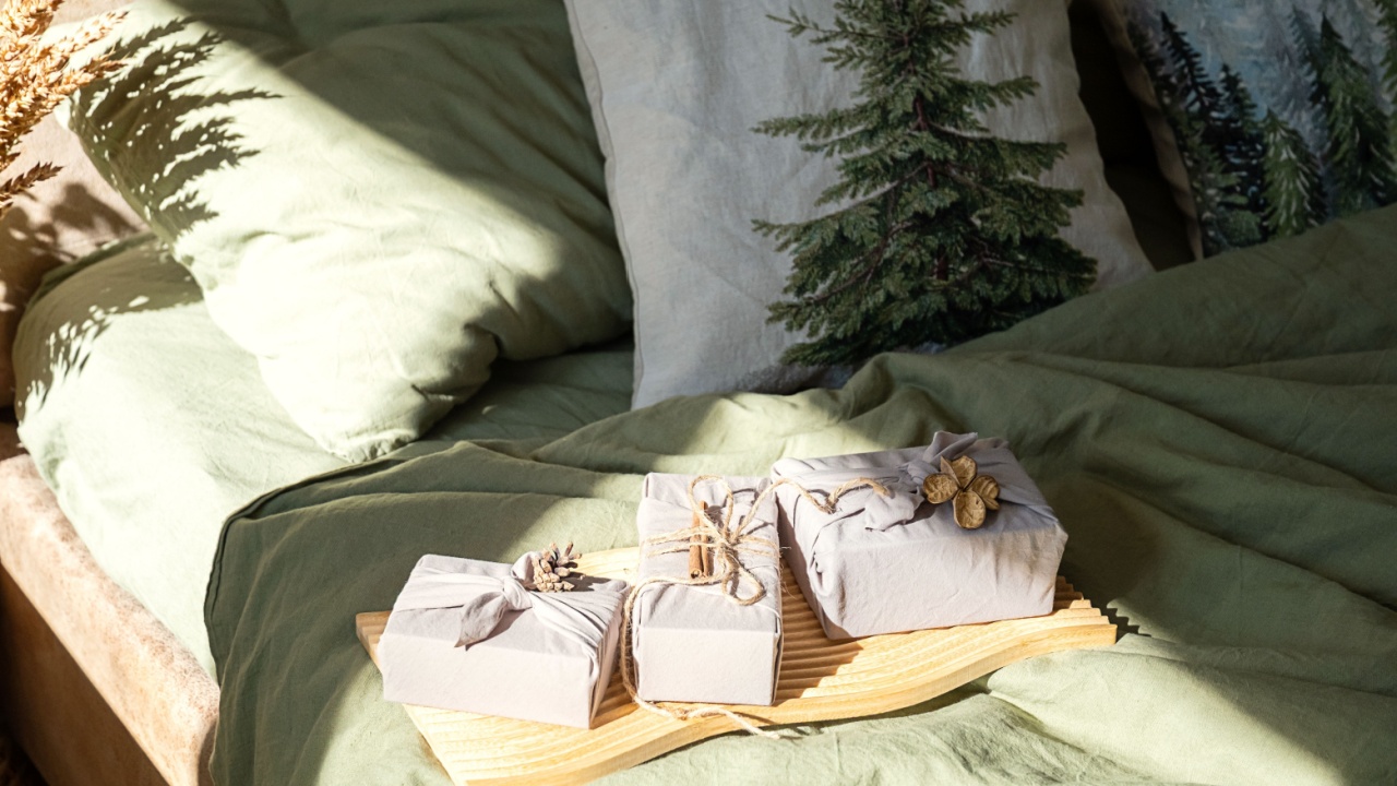 Three furoshiki wrapped gifts on wooden tray atop muted green bed with holiday themed pillows.