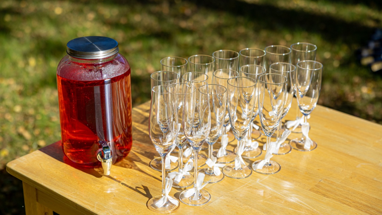 A red beverage dispenser with clear glasses tied with white ribbons stands on a wooden table outdoors, ready for guests at a bright festive event