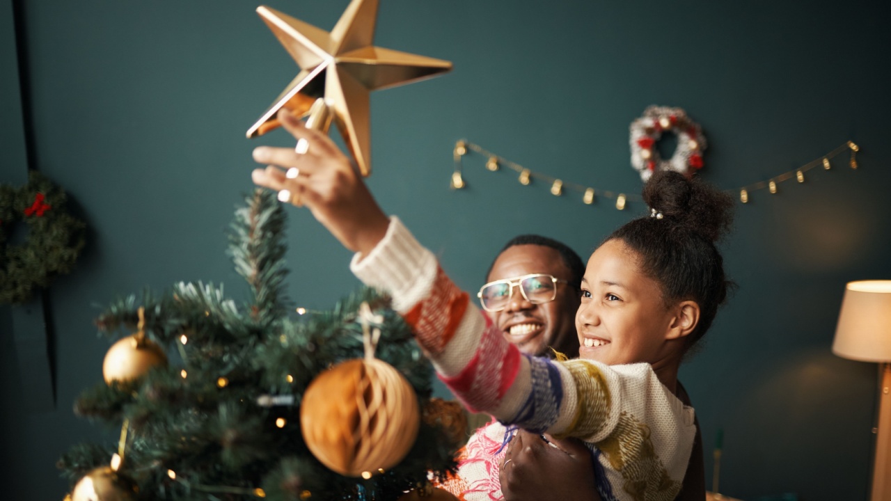 Black man lifting smiling Black girl reaching to place star on Christmas tree, both looking at decorations, festive ornaments and lights visible in background, joyful holiday moment