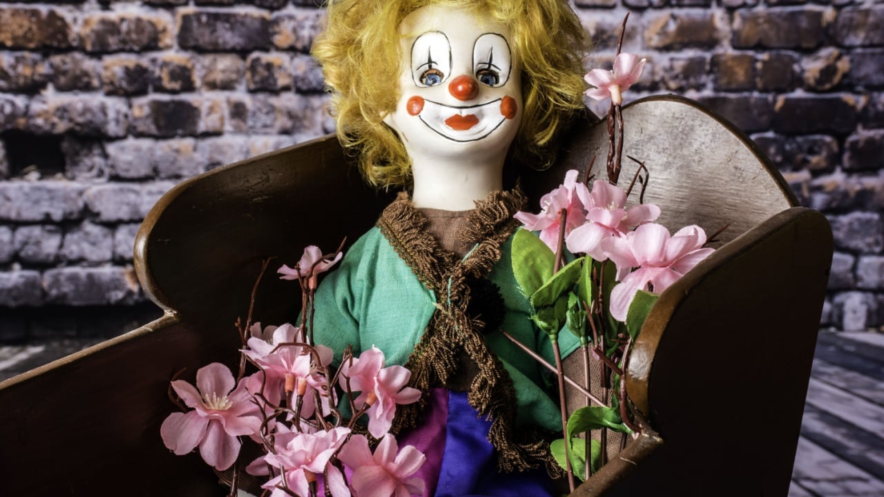 porcelain clown doll holding pink flowers in antique wood cradle with brick wall background