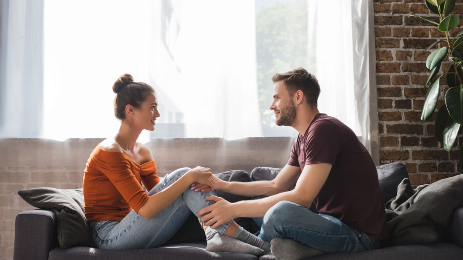 Side-view-of-young-couple-talking-while-sitting-on-sofa-and-holding-hands