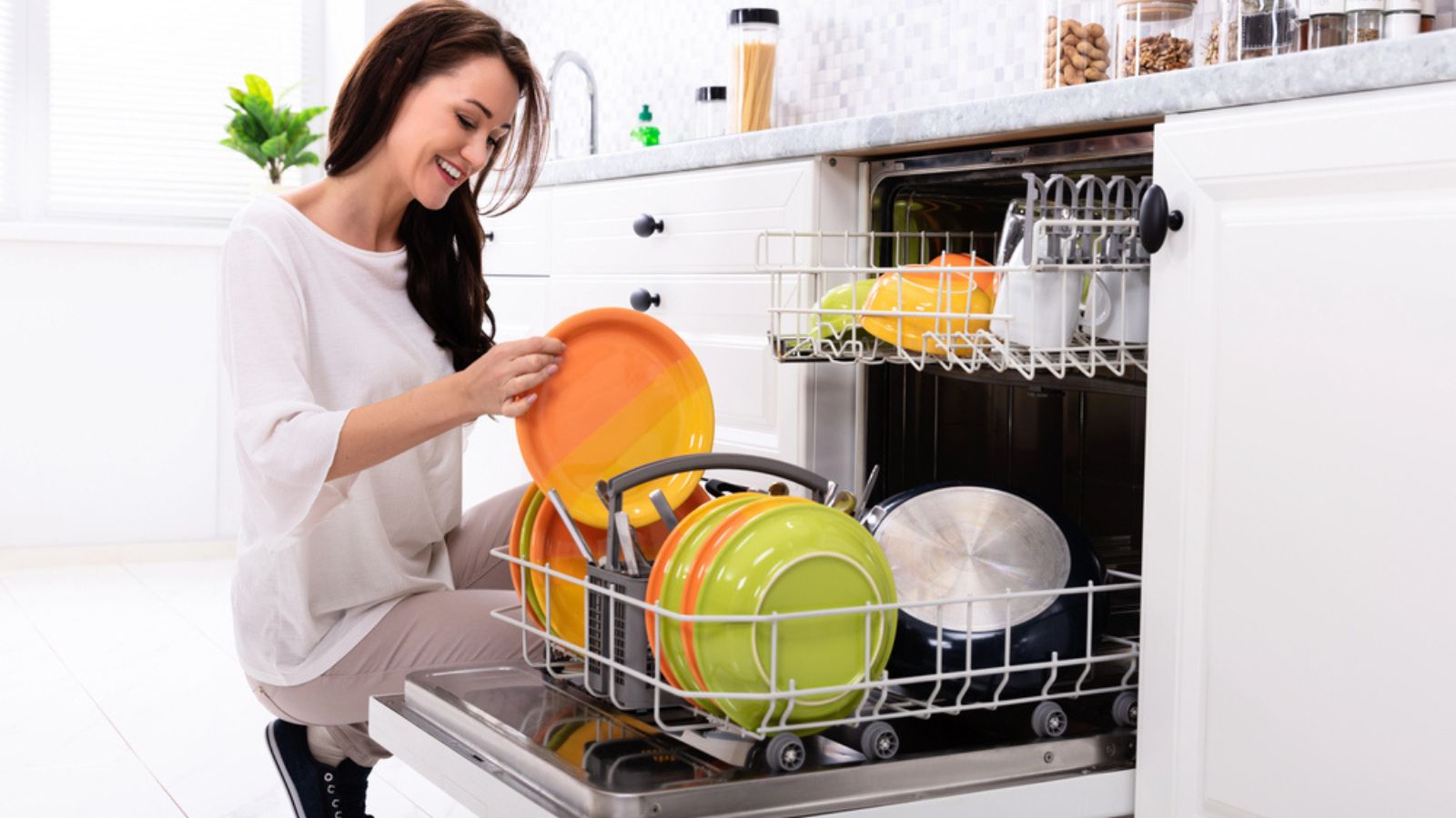 Smiling Young Woman Arranging Plates In Dishwasher At Home