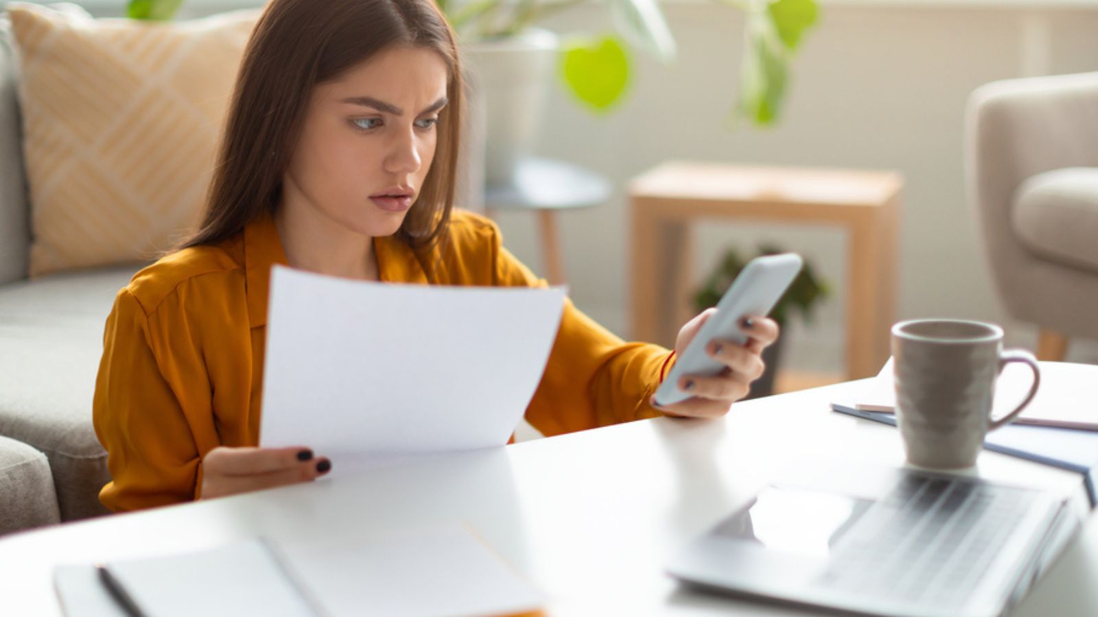 Stressed young woman with document using mobile phone and laptop