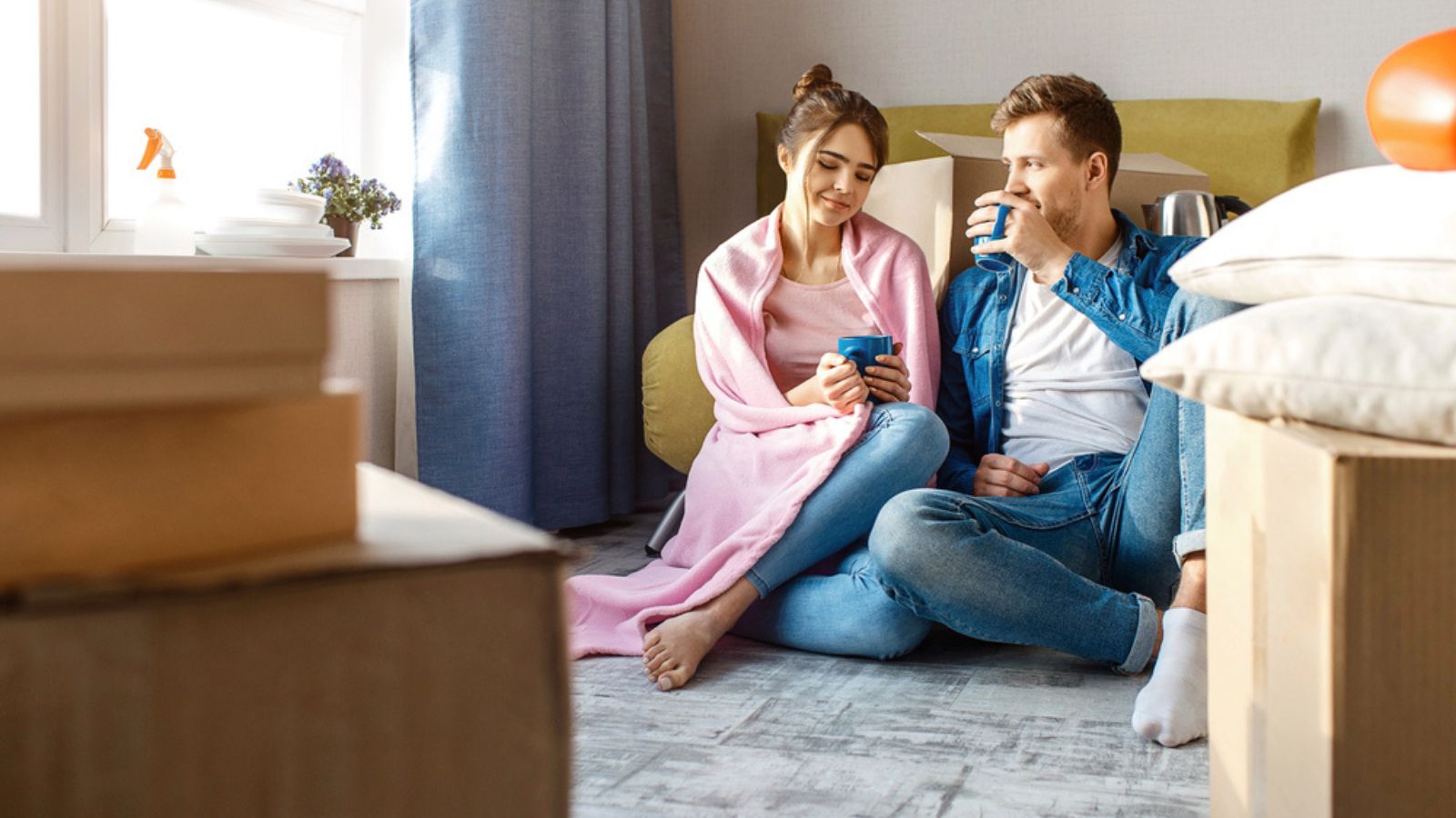 Young-family-couple-bought-or-rented-their-first-small-apartment.-They-sit-in-floor-and-drink-from-blue-cups.-Chilling-and-having-rest-after-unpacking