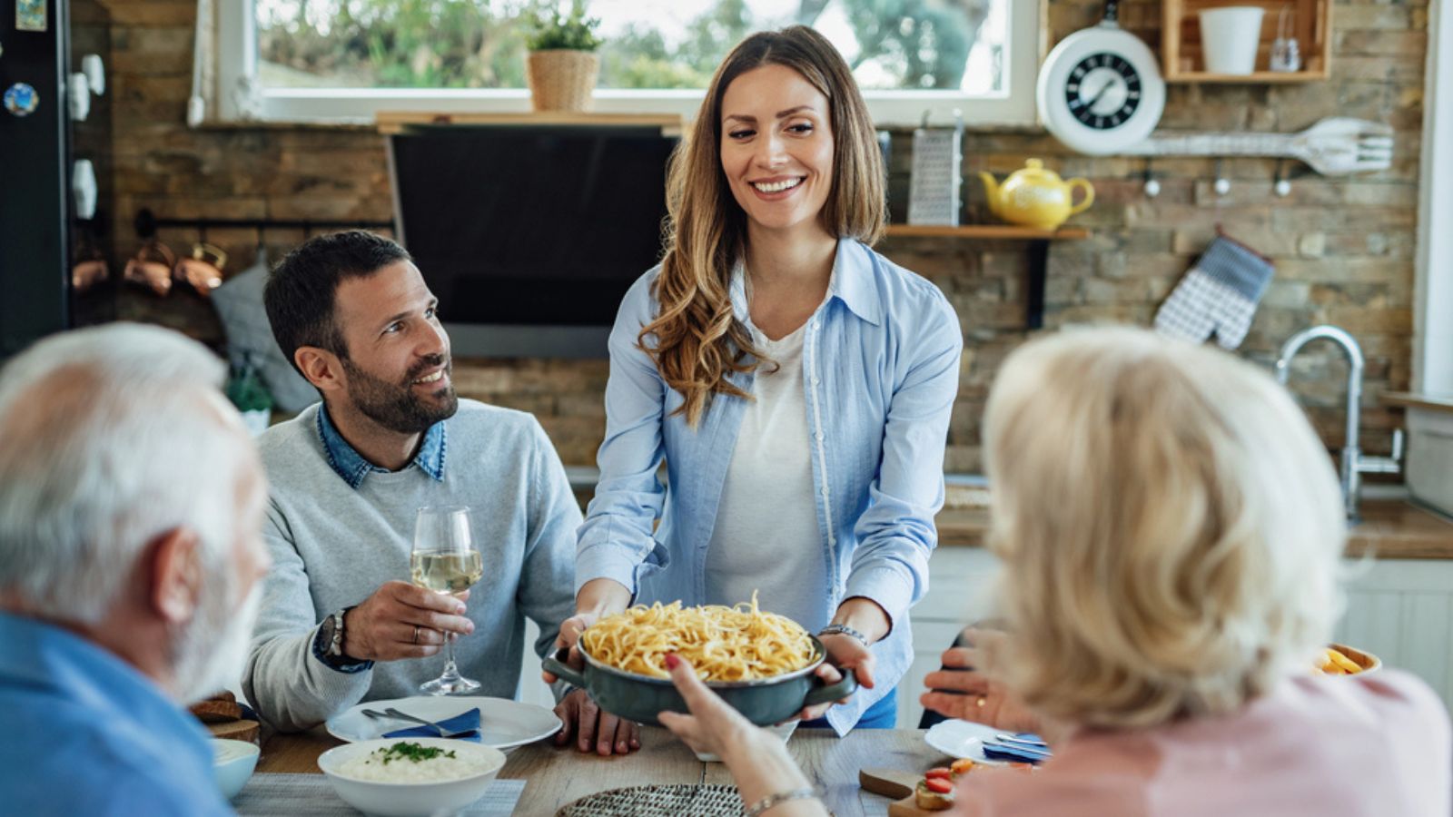 Young-happy-couple-and-their-mature-parents-having-lunch-together.-Focus-is-on-woman-bringing-food-at-dining-table