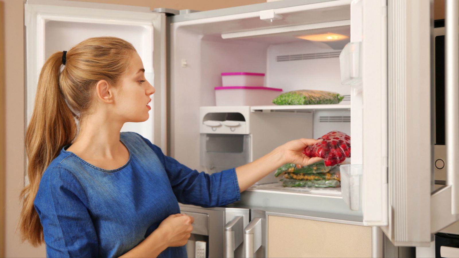 Young woman with frozen cherries near open refrigerator at home