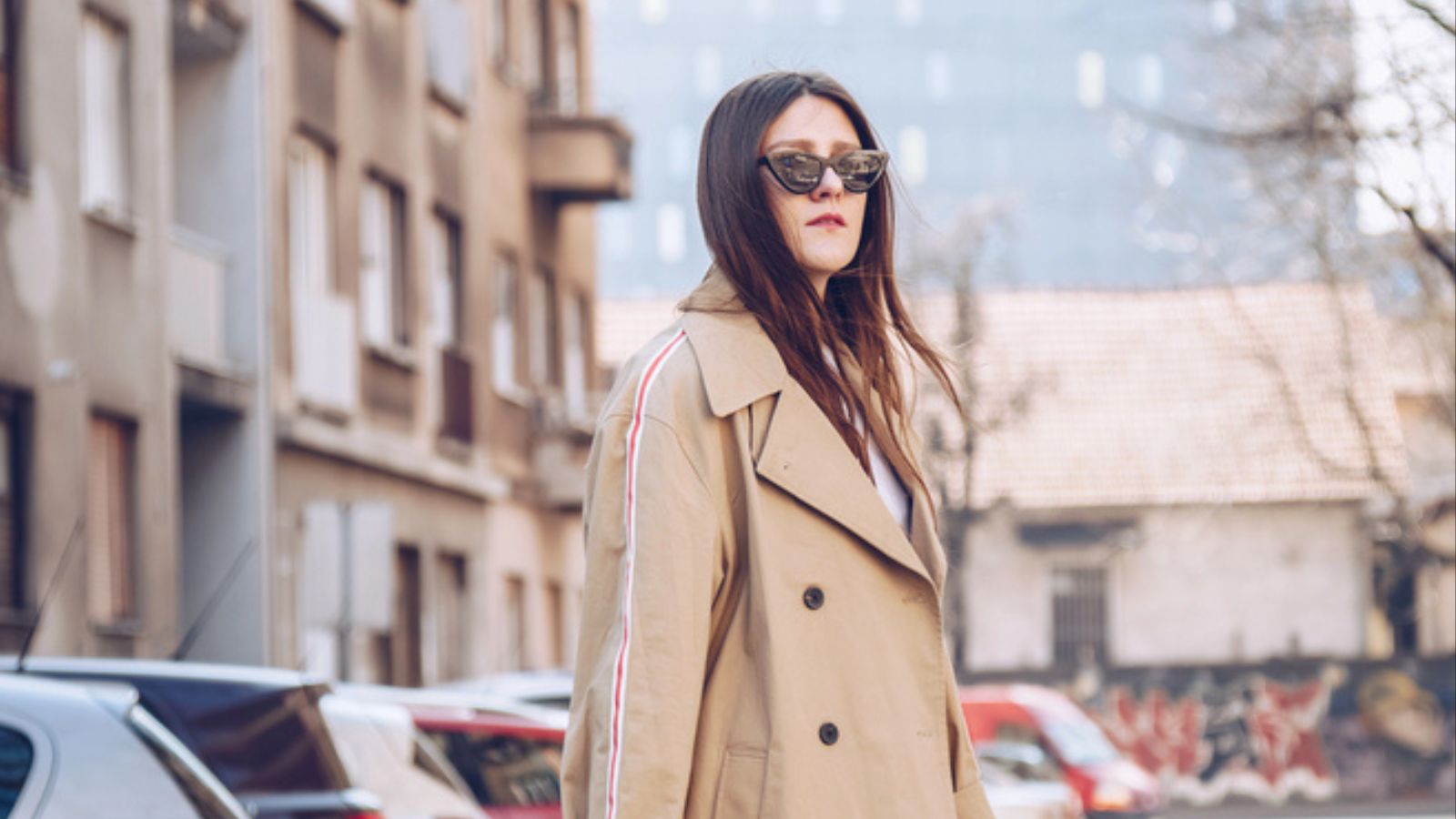 woman wearing a beige trench coat, denim jeans, ankle boots, cat eye sunglasses and a metallic handle brown tote bag. fashion outfit perfect for sunny spring day