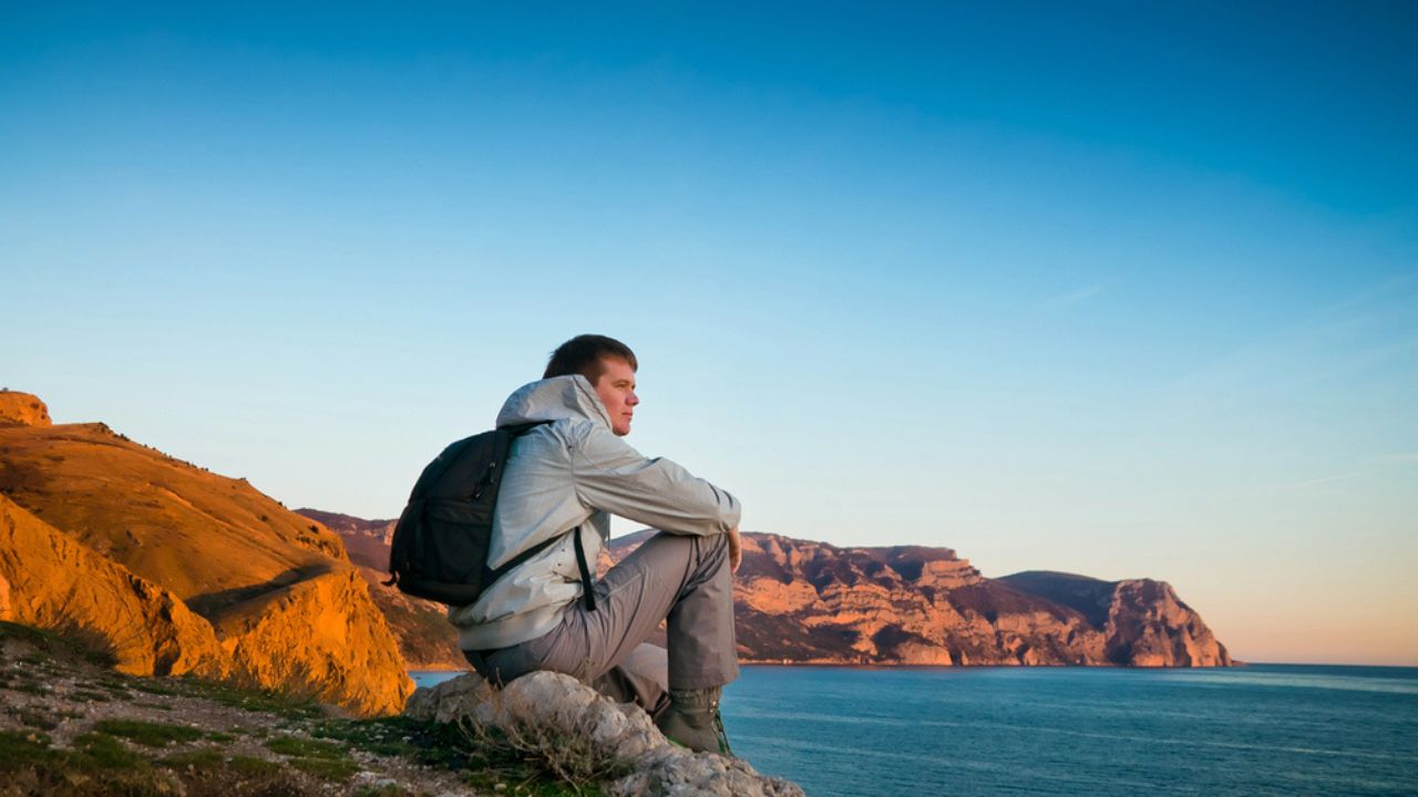 A young man sits on a hill and enjoys sea sunset