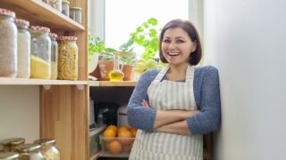 A woman in an apron stands smiling, next to her pantry