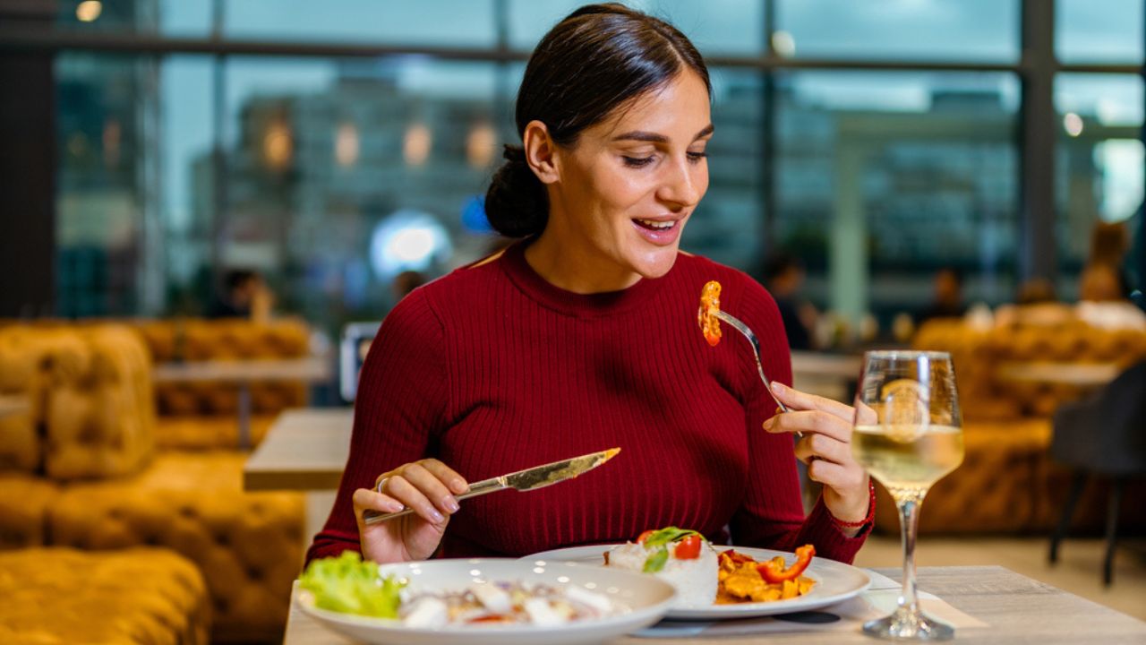 Attractive brunette in a red dress taking a bite of her lunch, eating pasta in the restaurant with a city view