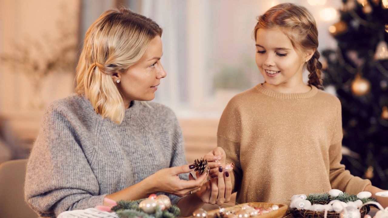 Attractive young blonde mother sitting at table and discussing natural decorations for Christmas wreath with daughter at home