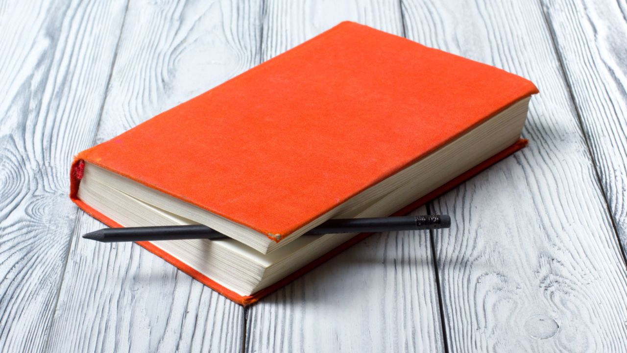 "Blank red hardcover book with black pencil on white wooden background.
