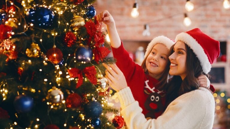 a mother holds a little girl as she places an ornament on a Christmas tree