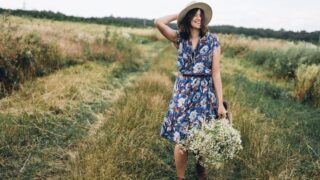 Happy-woman-in-blue-vintage-dress-and-hat-walking-with-white-wildflowers-in-straw-basket-in-meadow.-Beautiful-girl-with-white-daisies-bouquet-in-field