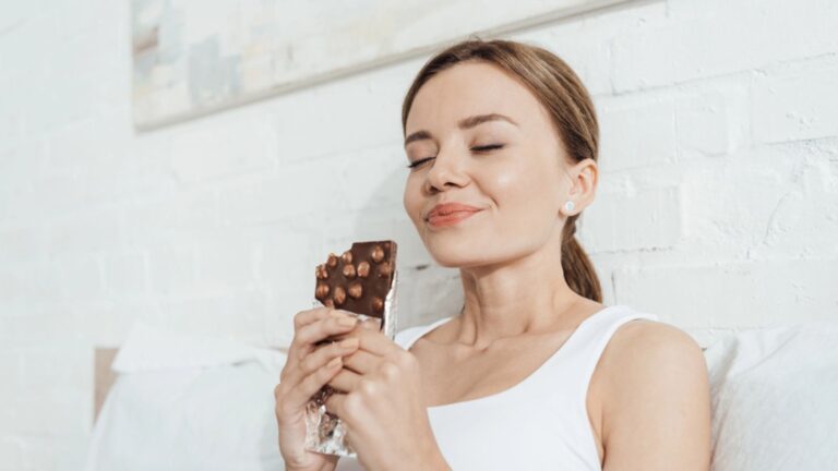 Low angle view of smiling young woman eating chocolate with nuts with closed eyes in bed