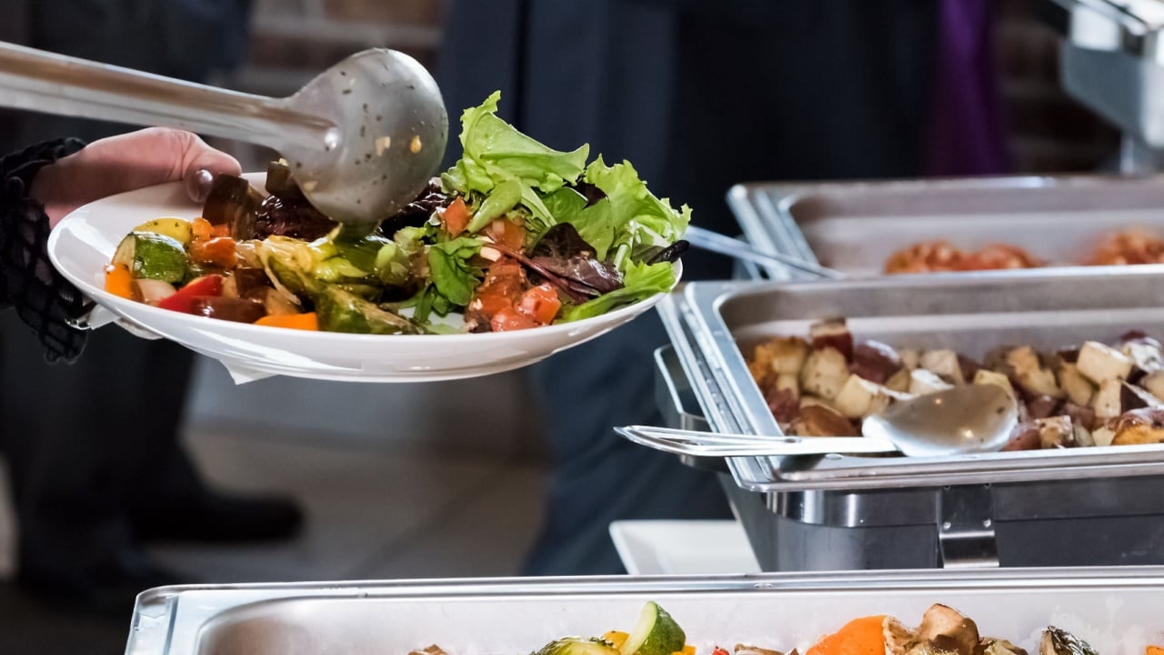 Fresh grilled vegetables in hot buffet tray with closeup of woman using tongs to serve food to plate in banquet, wedding, or restaurant inside