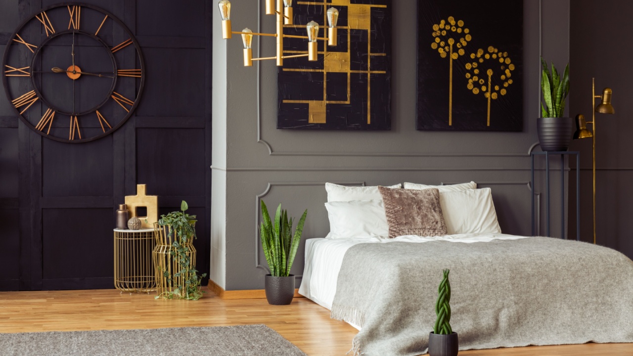 Real photo of a spacious bedroom interior with grey walls, big clock, paintings, plants, bed and golden accents