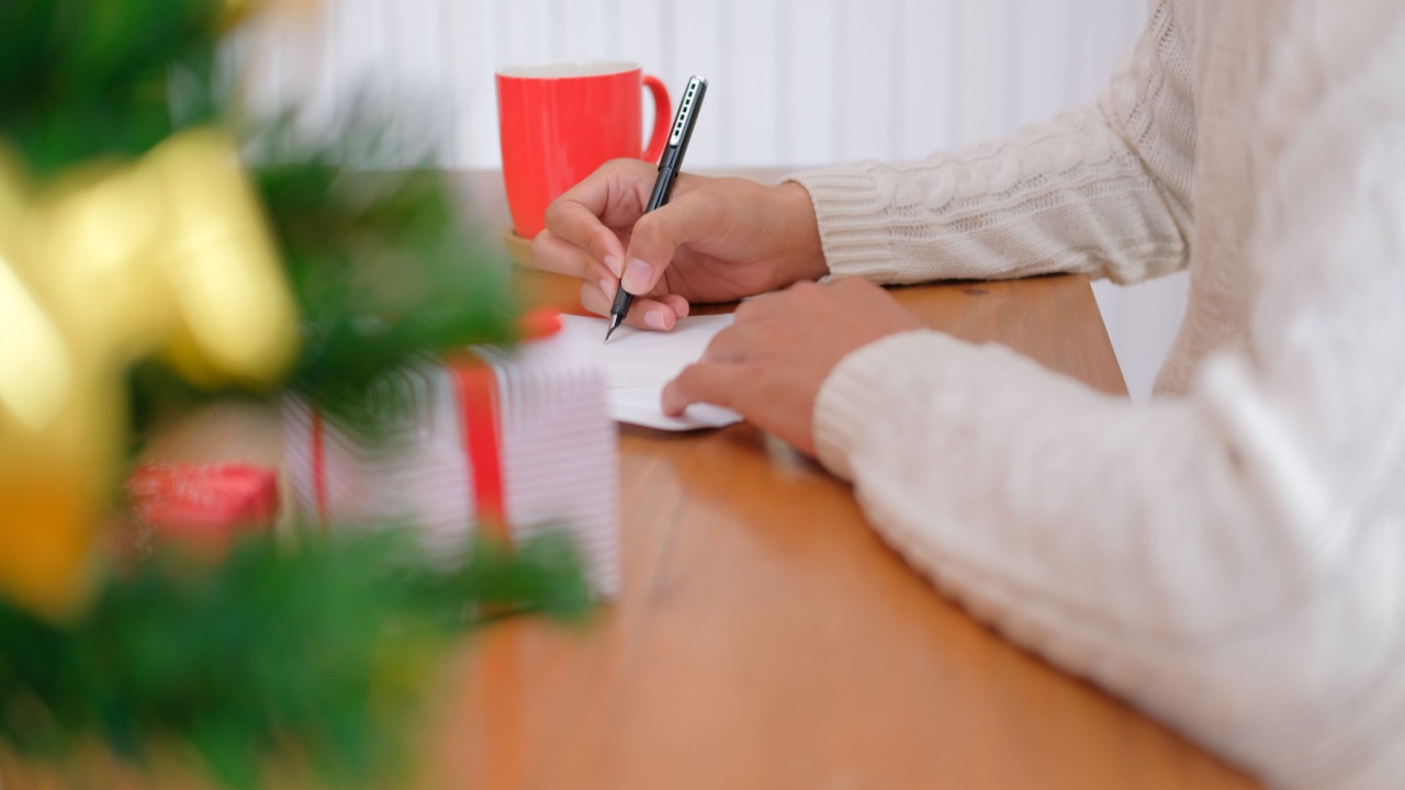 man wearing cream sweater writing christmas letter greeting card holiday wishes with xmas decoration.