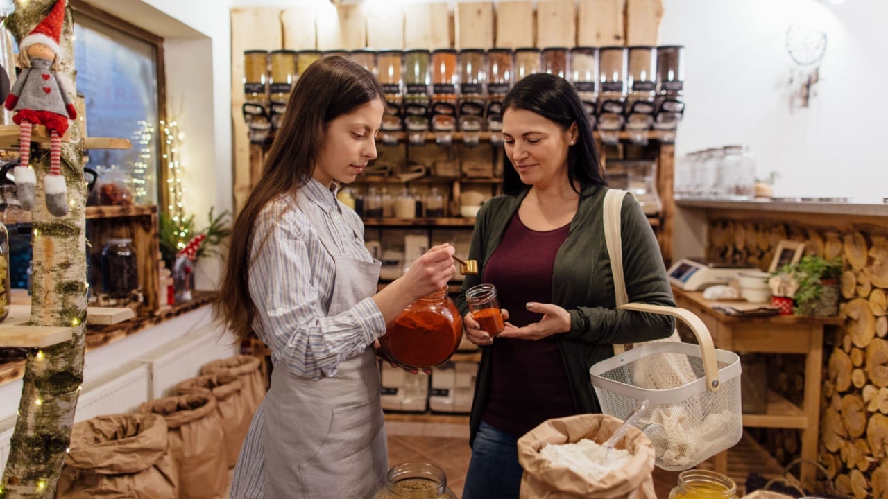 Woman buying herbs and spices in plastic free shop. Shop owner scooping red paprika in glass jar for customer in zero waste grocery store.