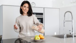 Image of brunette woman 30s making breakfast with oatmeal and fruits while standing in modern kitchen at home