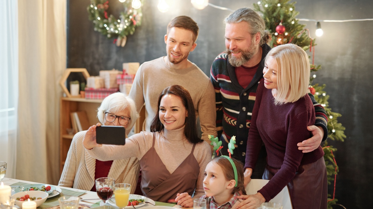 Cheerful affectionate family of six looking at smartphone camera while making selfie by served festive table at home