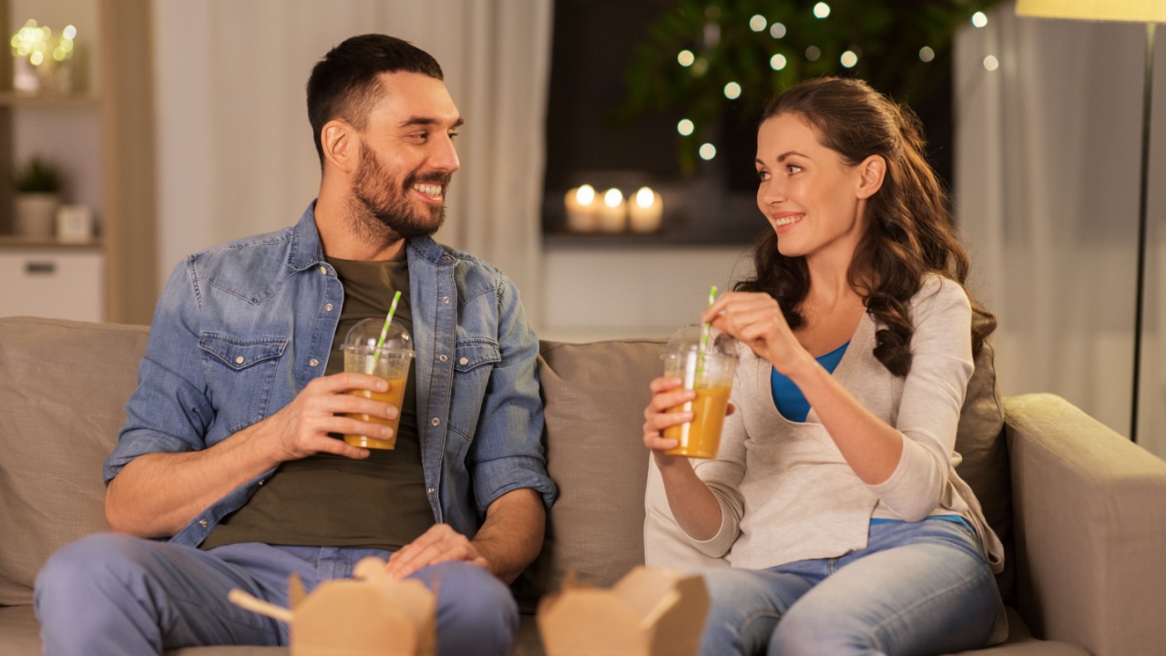 drinks and people concept - happy couple drinking takeaway juice from plastic cups with paper straws at home in evening