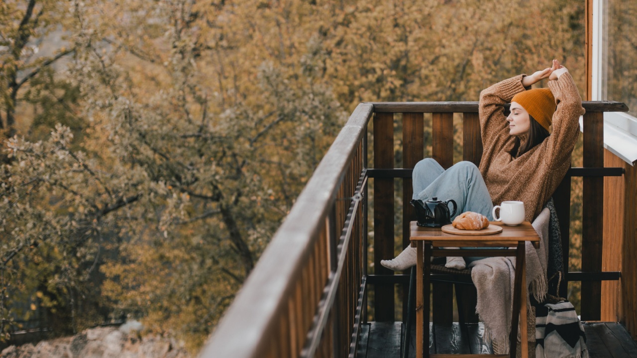 Young woman in knitted sweater and hat drinking tea and eating fresh croissants on cozy balcony of a wooden country house on autumn day.