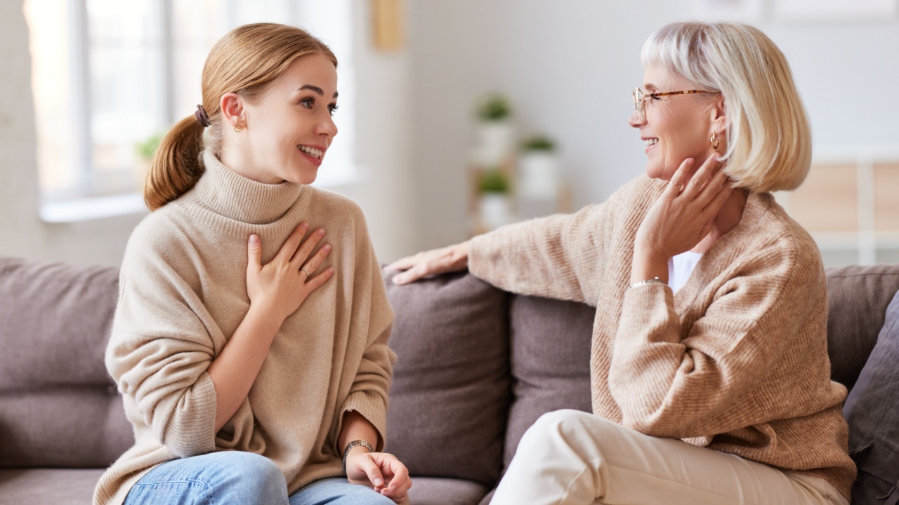 Young woman smiling and talking with senior mother while sitting on comfortable sofa at home together