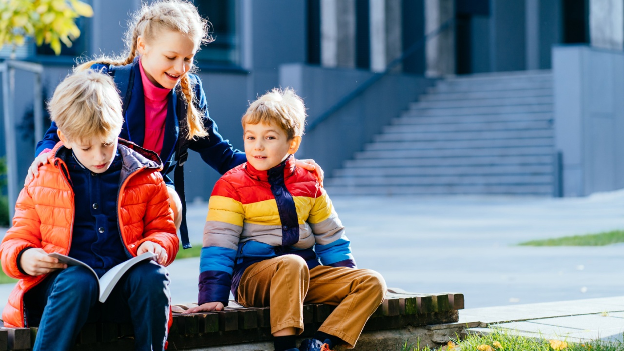 Group of three children reading from notebook all together. School kids revising notes for exams sitting on the bench outside of elementary school. School boys and girl studying for classwork