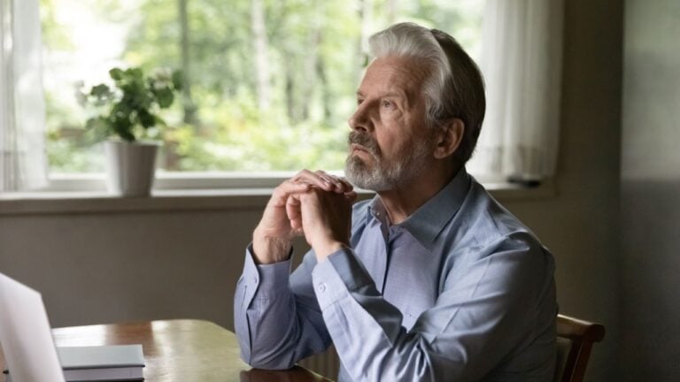 thinking mature grey haired 60s - 70s aged man sitting at table with laptop at home, looking away lost focus. Thoughtful retired OAP working at computer from home, thinking over retirement during break