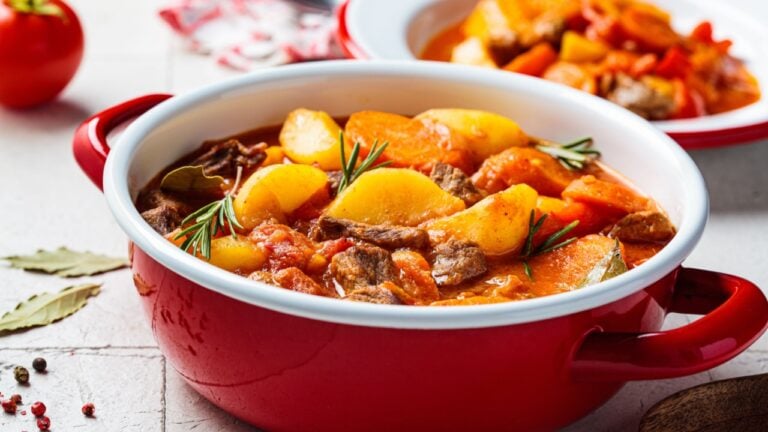 Beef stew with potatoes and carrots in tomato sauce in red pot, gray background. Slow cooking concept.