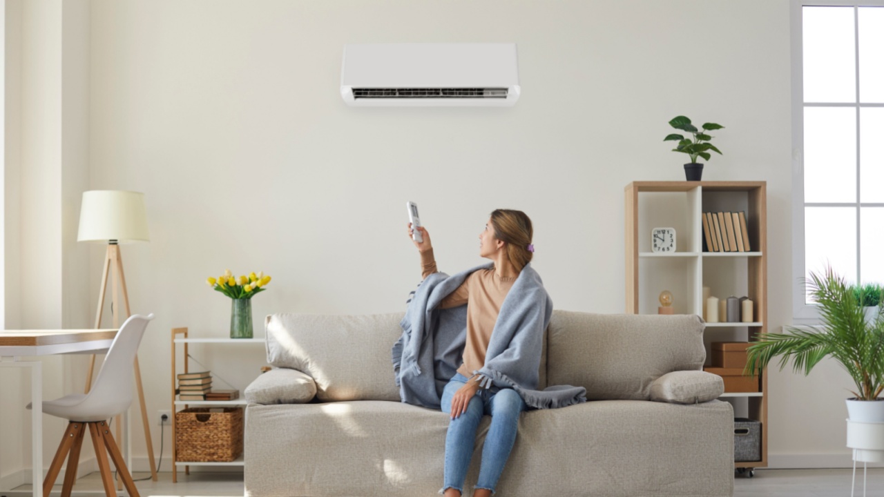 Woman who's sitting on sofa under warm plaid in living room switches off her air conditioner on wall. Young girl adjusting modern AC system, regulating temperature and enjoying cool fresh air at home