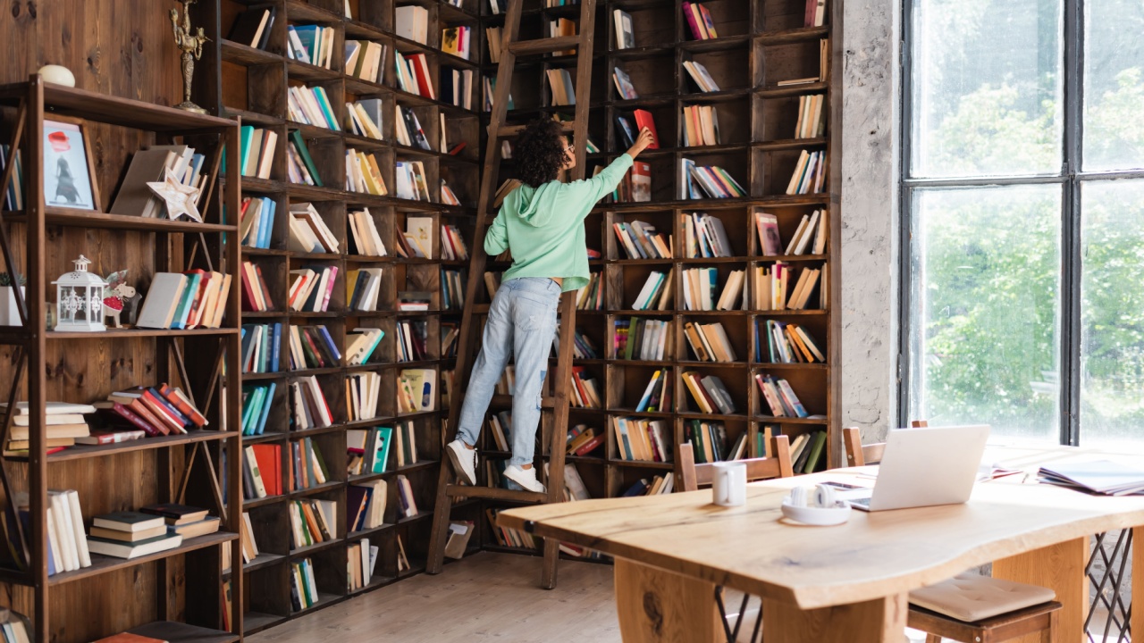 african american student standing on wooden ladder while reaching book near blurred gadgets on desk