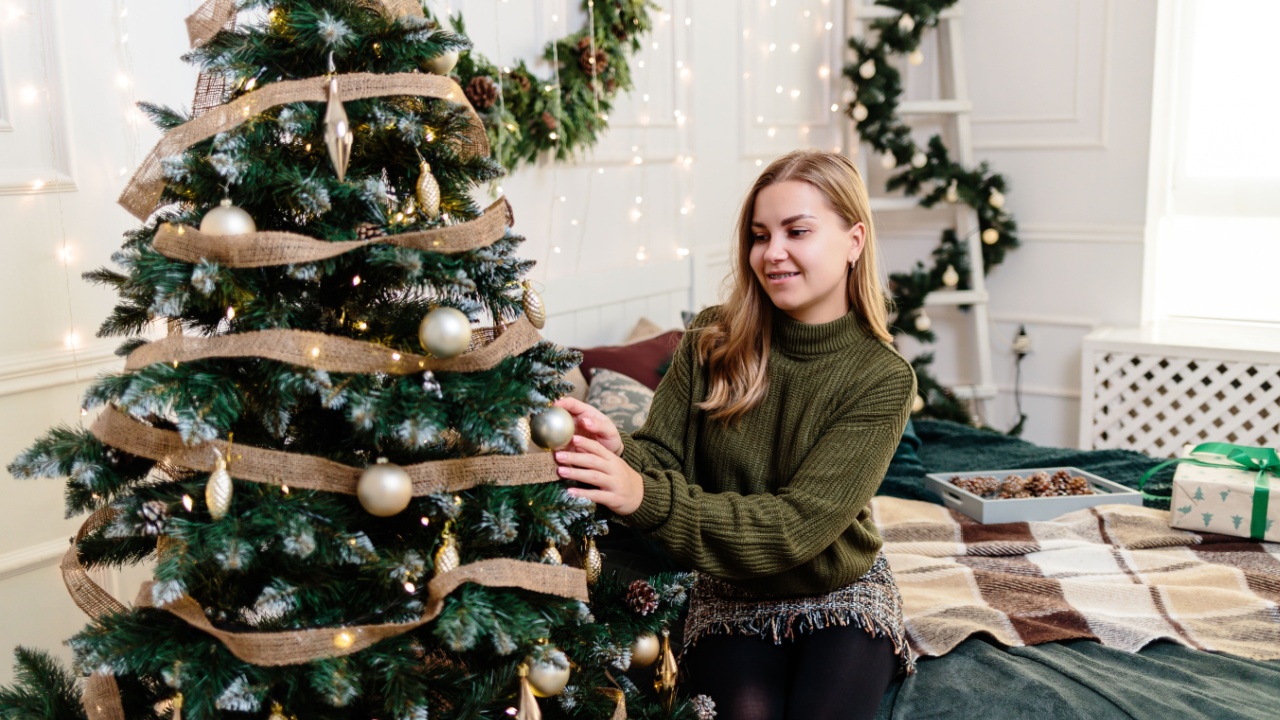 A beautiful young woman with blond hair sits near a Christmas tree and decorates with toys. New Years is soon. Christmas atmosphere in a cozy home