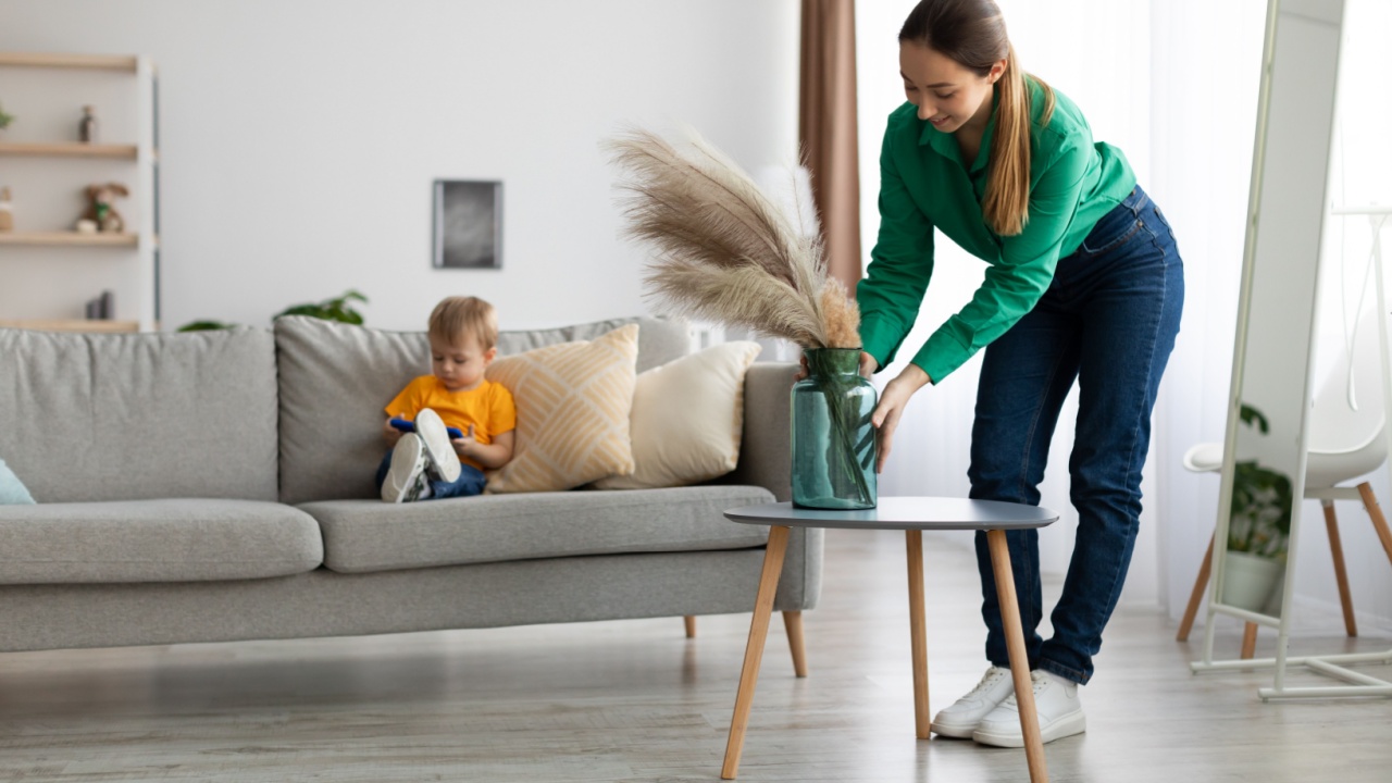 Casual young mother decorating living room and putting glass vase with dried flowers on tea table, while her little child sitting on sofa and watching cartoons on cellphone
