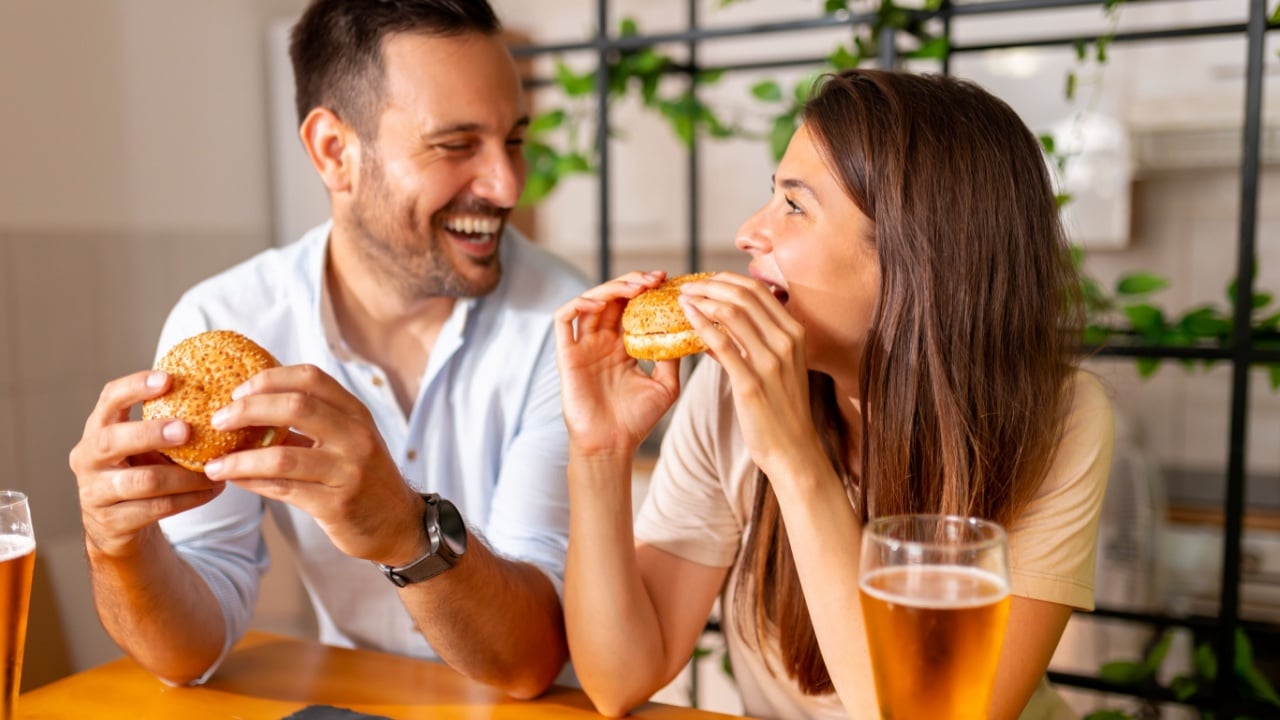 Beautiful young couple in love having fun spending leisure time together at home, eating burgers and drinking beer