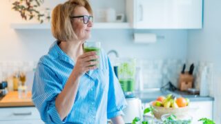 Smiling middle aged woman with just made glass of detox shake, green smoothie in the kitchen. Healthy dieting, eating, cooking. Natural anti aging methods