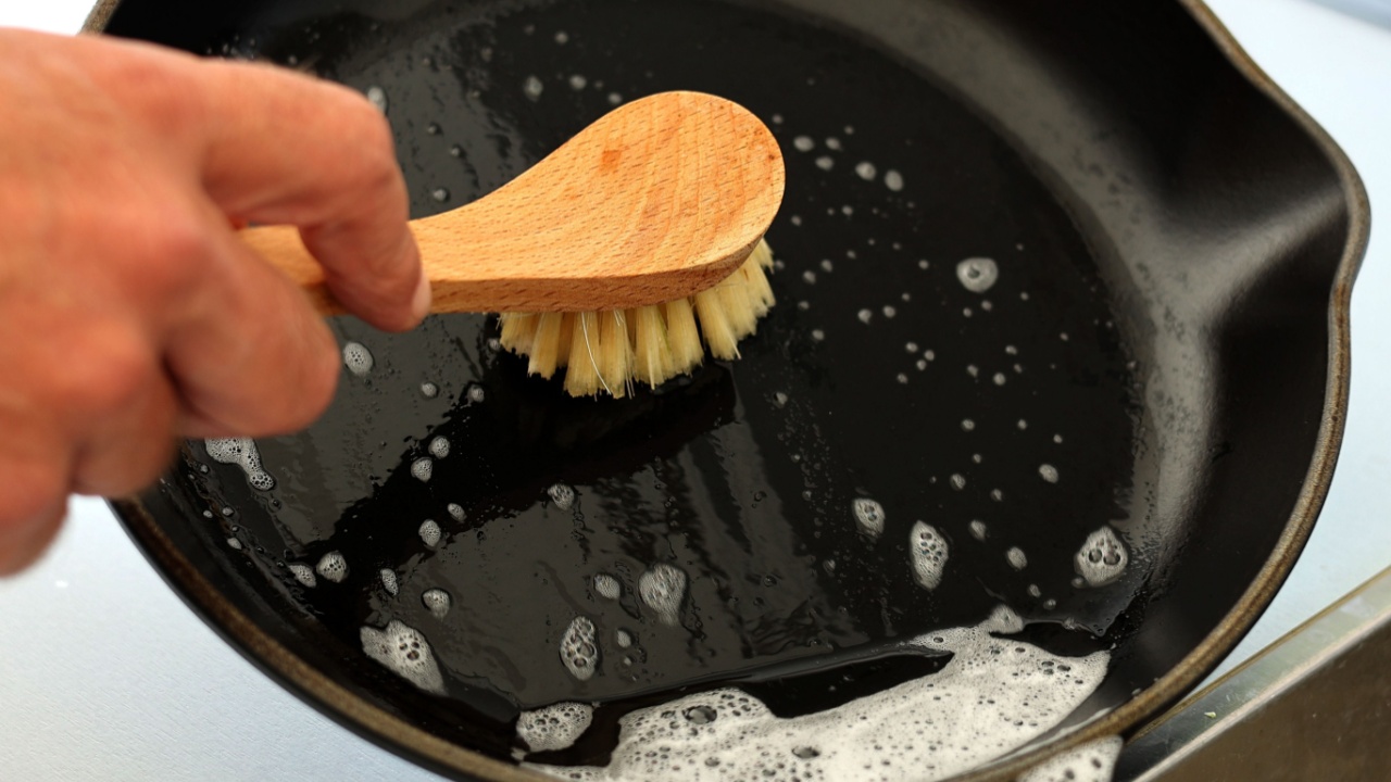 Close up A hand holding bamboo cleaning brush scrub and washing the dirty cast iron pan with a pattern of grease and oil stains.