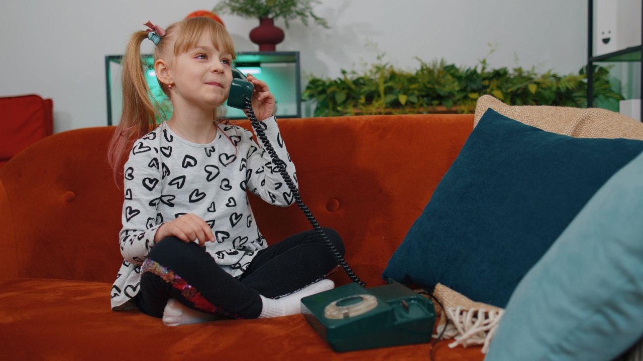 Smiling young little teen children girl making fun conversation call on landline retro telephone with friends, sitting on couch at home. Happy excited toddler child kid enjoying talking communication