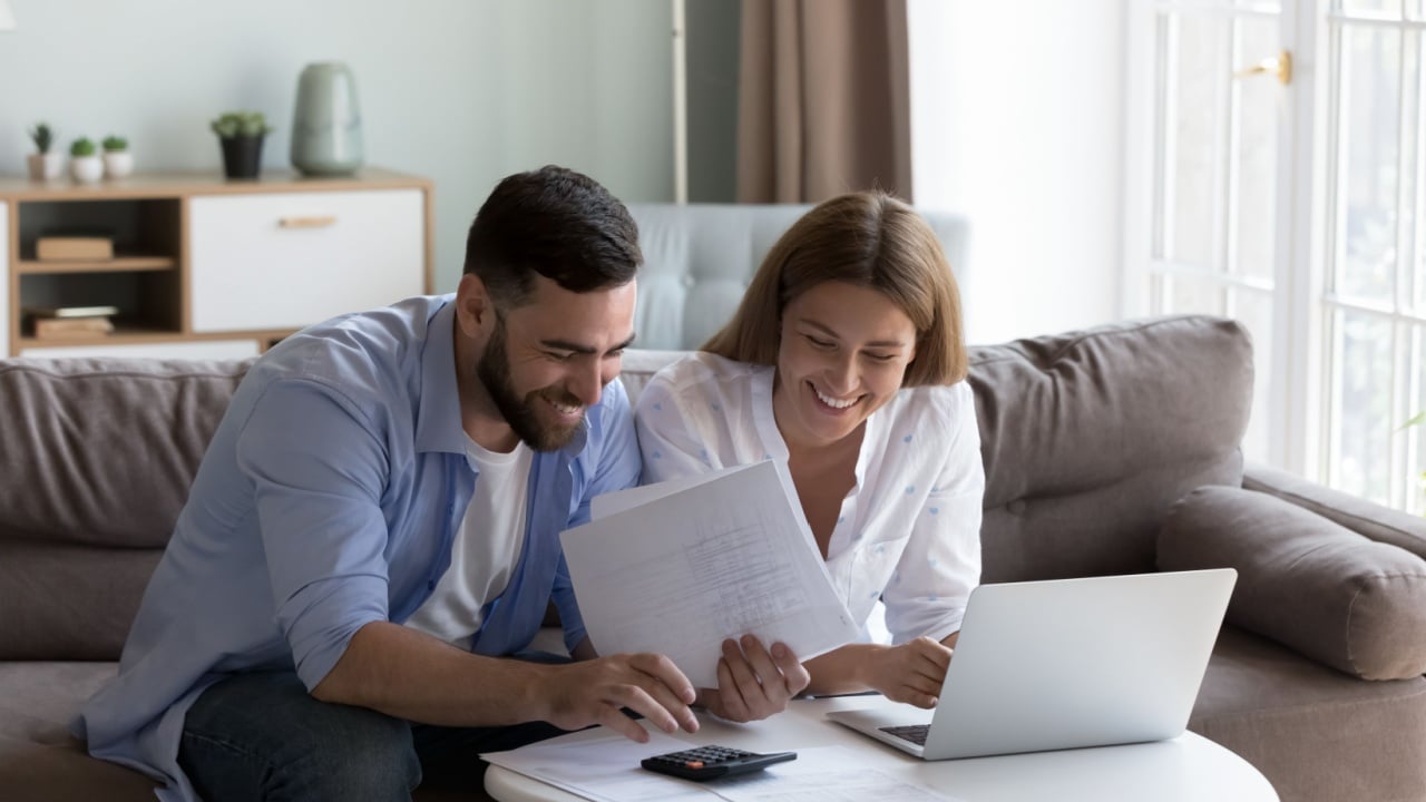 Cheerful young millennial husband and wife doing domestic paperwork, accounting job, reviewing paper bills, receipts at laptop computer, using calculator, paying mortgage, rent fees on internet
