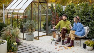 Young stylish couple hang out together, sitting with wine by the fire, spending evening time at cozy atmosphere in beautiful backyard with greenhouse on background