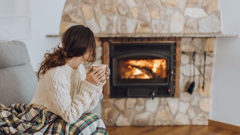 Young woman sitting at home by the fireplace with a hot tea or coffee mug and warming her hands, she is wearing white woollen sweater. Cold houses in Europe concept during energy and gas crisis.