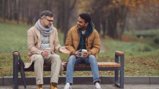Friends, a senior and a young man sit in the park on a bench and talk in the autumn park.