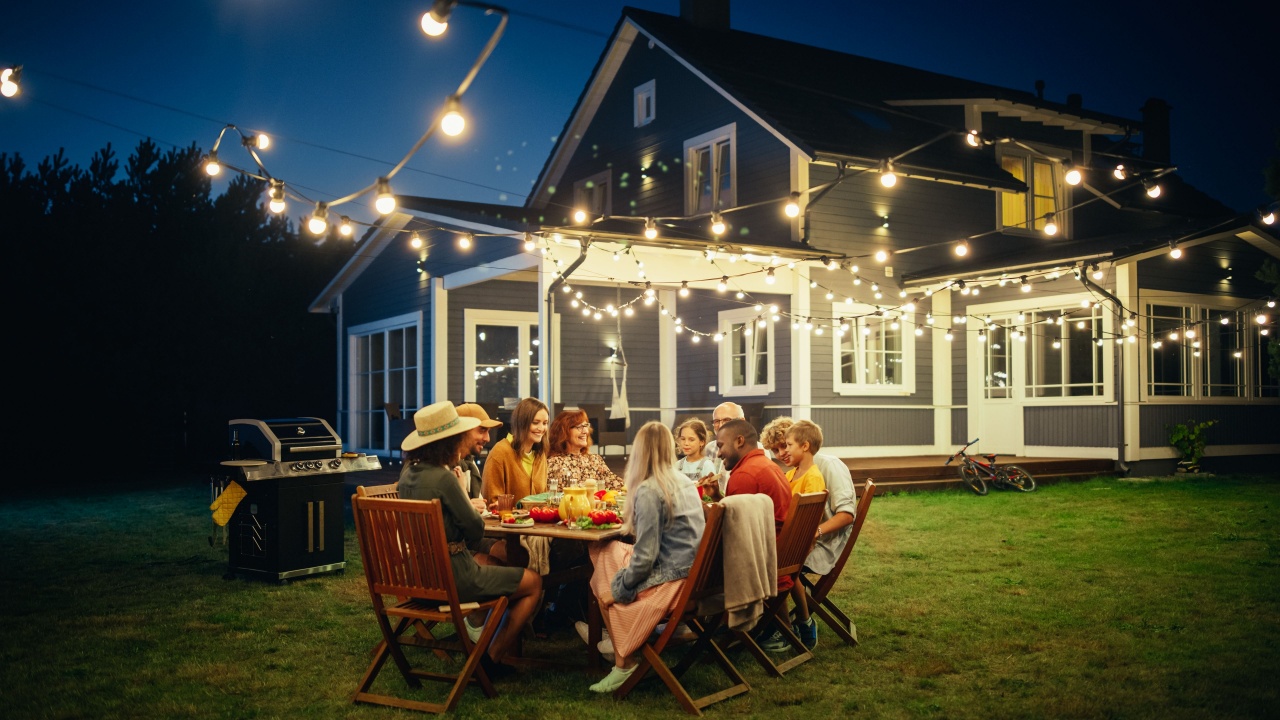 Parents, Children and Friends Gathered at a Barbecue Dinner Table Outside a Beautiful Home with Lights Decorations. Old and Young People Have Fun and Eat Food. Garden Party Celebration in a Backyard.