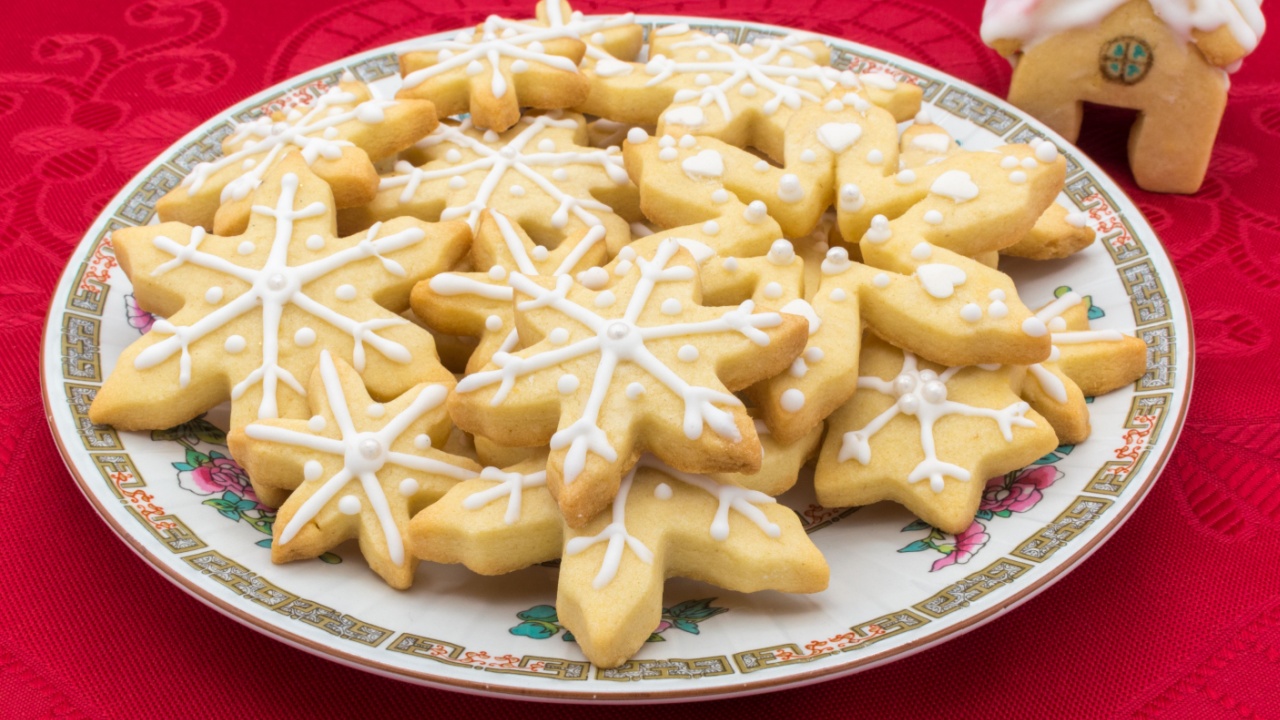 Plate with snowflake-shaped sugar icing Christmas cookies. Disposed on a red tablecloth with a house-shaped cookie as decoration.