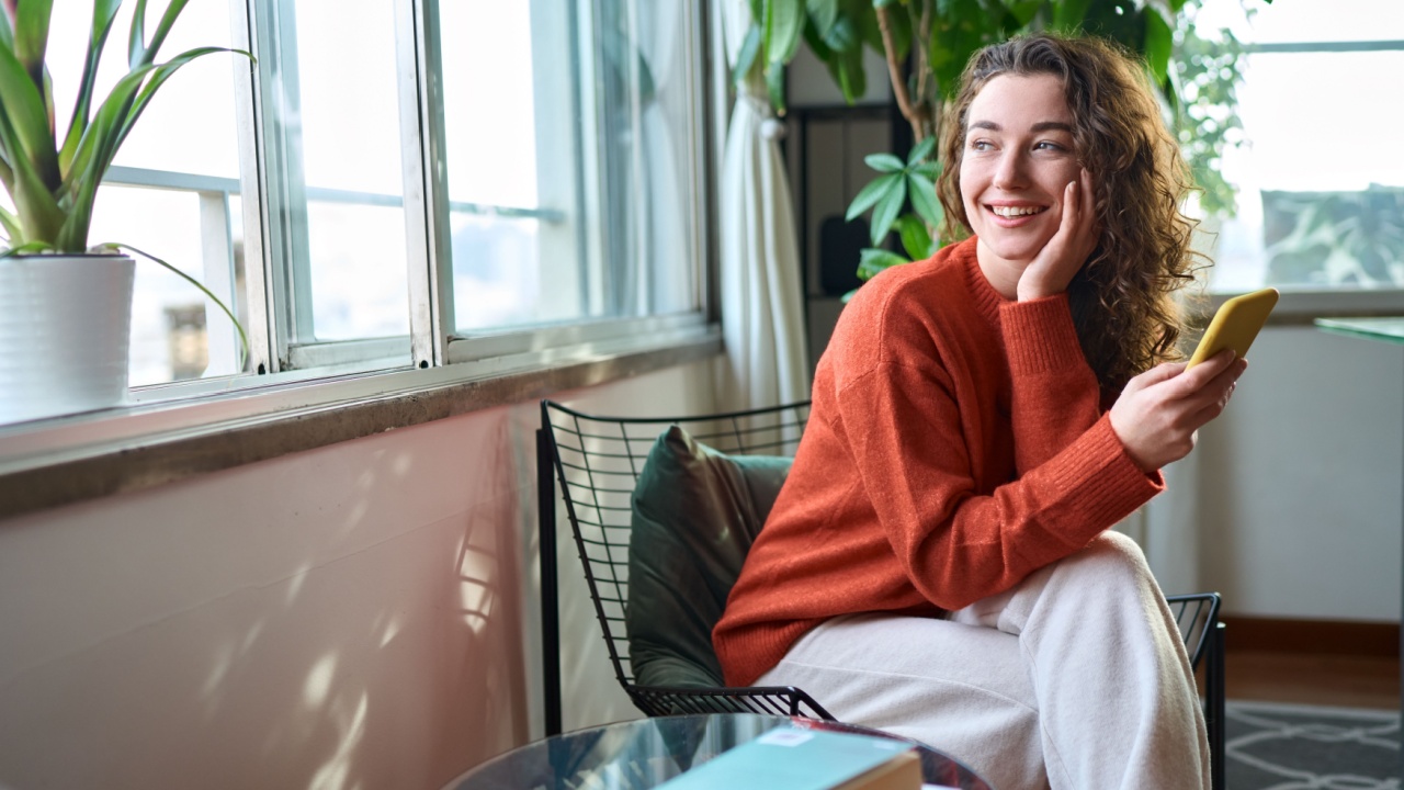 Happy young woman sitting on chair holding mobile phone using cellphone device, checking modern apps on smartphone, texting messages, browsing internet doing shopping relaxing at home.